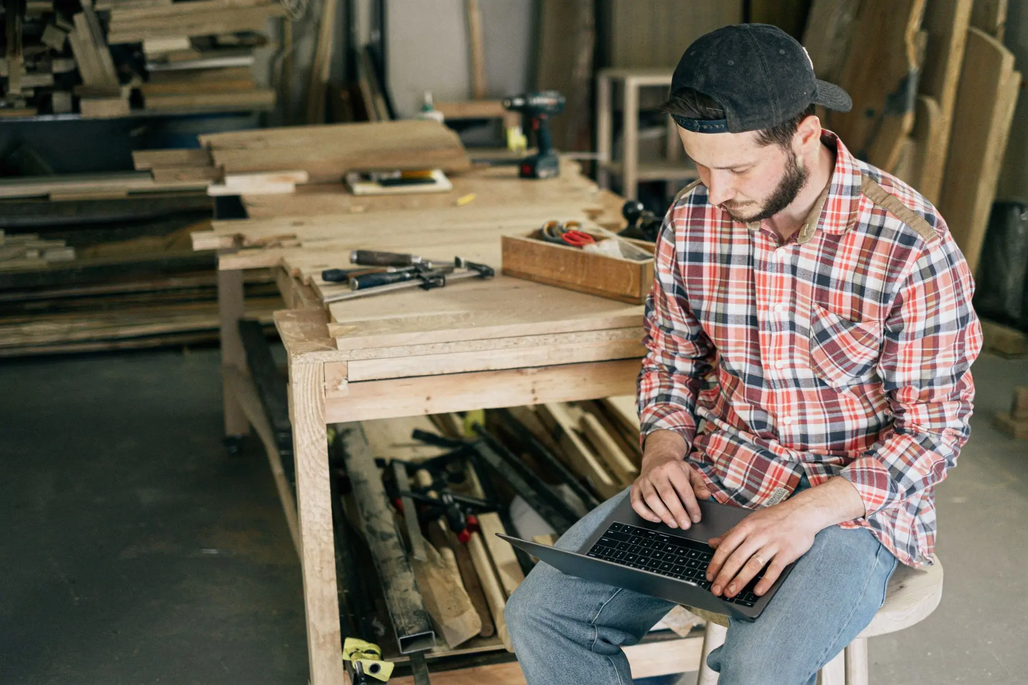 Joinery business owner sitting at his workbench checking an online register on a laptop, surrounded by timber offcuts and hand tools